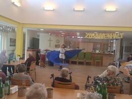 Eine Person mit einem großen blauen Umhang führt auf einer Bühne auf. Im Hintergrund stehen Stühle und Tische mit Gästen, die aufmerksam zuschauen. An der Wand hängen farbige Buchstaben, die 