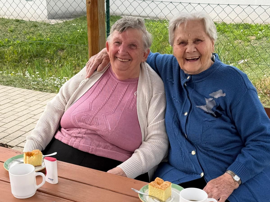 Zwei ältere Frauen sitzen an einem Tisch im Freien, lächeln und umarmen sich. Vor ihnen stehen Tassen und Stücke Kuchen auf Tellern. Im Hintergrund ist ein grüner Rasen und ein Zaun zu sehen.