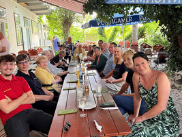Eine Gruppe von Menschen sitzt an einem langen Tisch im Freien, umgeben von Bäumen und Sonnenschirmen. Sie lächeln und posieren für das Foto, während Teller und Gläser auf dem Tisch stehen. Das Ambiente wirkt gemütlich und einladend.