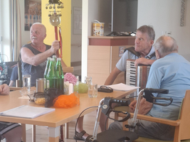 Zwei Männer zeigen auf eine Spielzeugfigur, während einer ein Akkordeon spielt. Um sie herum sitzen ältere Menschen, einige mit Gehhilfen. Auf dem Tisch stehen Getränke und Snacks. Der Raum wirkt einladend und lebhaft, typische Aktivitäten in einer Seniorenresidenz.