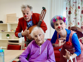 Clowns unterhalten eine ältere Frau im Rollstuhl in einem freundlichen Raum.
