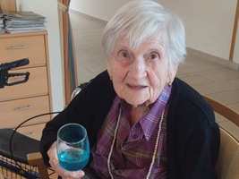 Ältere Frau sitzt am Tisch und hält ein Glas mit blauem Getränk.