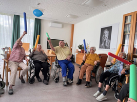 Ältere Menschen in Rollstühlen spielen mit bunten Schaumstoffstäben und einem Luftballon.