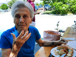 Ältere Frau in blauer Bluse sitzt am Tisch mit einem Teller Essen.