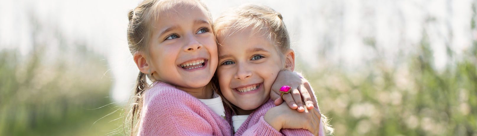 Zwei lächelnde Mädchen umarmen sich in einer blühenden Landschaft. Beide tragen rosa Pullover und haben blonde Haare. Der Hintergrund zeigt eine Reihe von Bäumen, die mit Blüten bedeckt sind. Die Stimmung ist freundlich und fröhlich.