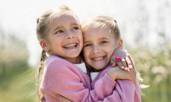 Zwei lächelnde Mädchen umarmen sich in einer blühenden Landschaft. Beide tragen rosa Pullover und haben blonde Haare. Der Hintergrund zeigt eine Reihe von Bäumen, die mit Blüten bedeckt sind. Die Stimmung ist freundlich und fröhlich.