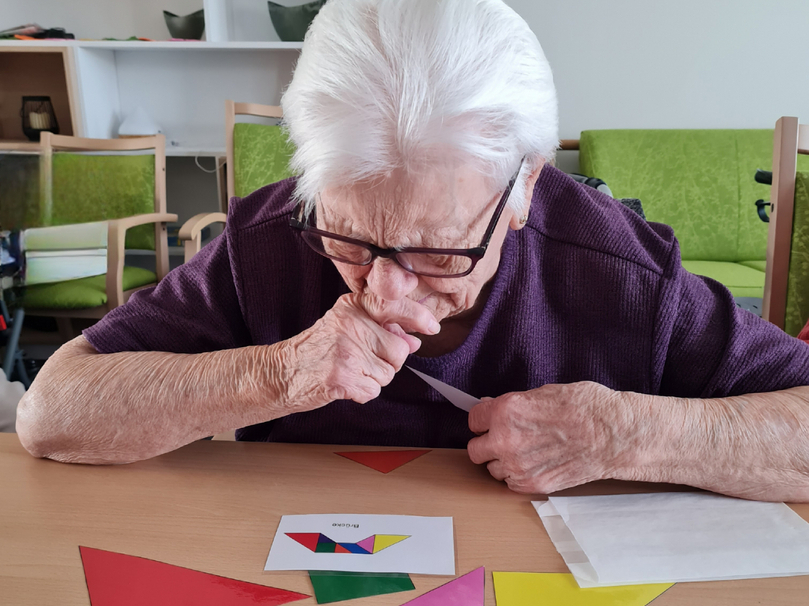 Eine ältere Frau mit Brille sitzt an einem Tisch und konzentriert sich auf bunte geometrische Formen, die vor ihr liegen. Sie hält einen Gegenstand in der Hand und betrachtet eine Karte, während sie an ihre Lippen denkt. Im Hintergrund sind Stühle und eine grüne Couch sichtbar.