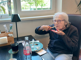 Eine ältere Frau sitzt an einem Tisch und genießt ihr Essen. Sie hält einen Löffel in der Hand und hat einen Teller mit Resten vor sich. Auf dem Tisch stehen Getränke und Besteck. Im Hintergrund sind Pflanzen und Fenster sichtbar.