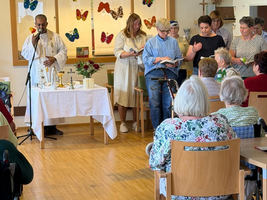 Eine Gruppe von Menschen führt eine Zeremonie in einem Raum durch, der mit Schmetterlingen dekoriert ist. Ein Mann mit Mikrofon und weißem Gewand steht an einem Tisch mit Blumen und Kerzen. Senioren sitzen in Stühlen und beobachten die Veranstaltung.