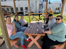 Eine Gruppe von sechs Personen sitzt an einem Holztisch unter einem Pavillon. Es ist sonnig, und im Hintergrund sind weitere Menschen und Pflanzen sichtbar. Einige Personen haben Getränke vor sich. Alle scheinen sich zu unterhalten und zu genießen.