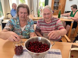 Zwei ältere Frauen sitzen an einem Tisch und füllen Einmachgläser mit Kirschen aus einer großen Schüssel. Im Hintergrund sind weitere Personen zu sehen, die an einem Tisch sitzen. Die Szene vermittelt eine Atmosphäre des gemeinsamen Arbeitens und der Geselligkeit.