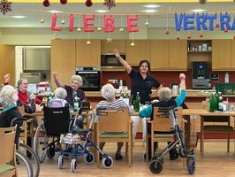 Eine Gruppe älterer Frauen hebt die Hände in einem Gemeinschaftsraum.