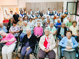 Eine Gruppe älterer Menschen sitzt in einem Raum, viele in Rollstühlen. Im Hintergrund stehen Musiker und singen. Die Stimmung ist freundlich und lebhaft, es gibt Lächeln und einige Teilnehmer winken. Der Raum ist hell und einladend gestaltet.