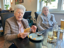 Zwei ältere Frauen sitzen an einem Tisch in einem Restaurant. Die erste Frau hält ein Stück Essen in der Hand und lächelt, während die zweite Frau daneben sitzt. Im Hintergrund sind andere Gäste und große Fenster zu sehen, die viel Tageslicht hereinlassen.