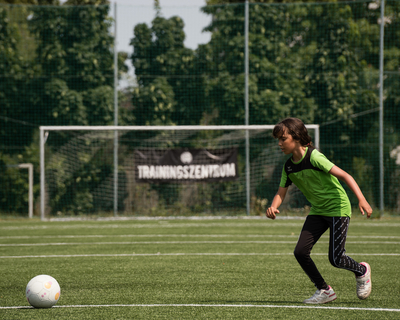 Fußballfeld mit Tor im Hintergrund, davor Mädchen in grünem Sportshirt lauft von rechter Seite des Bildes auf Fußball links zu