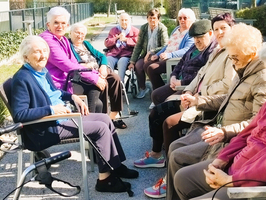Eine Gruppe von älteren Frauen sitzt auf einer Terrasse in einem sonnigen Ambiente. Sie sind in entspannter Unterhaltung vertieft, einige nutzen Gehwagen. Die Frauen tragen verschiedene Kleidung, die die frühlingshafte Atmosphäre widerspiegelt.