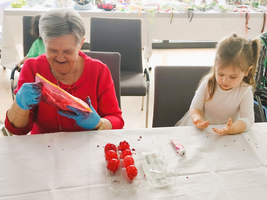 Eine ältere Frau in rotem Oberteil und blauen Handschuhen hält einen Beutel mit roter Farbschicht und lächelt, während ein kleines Mädchen in einem hellen Oberteil an einem Tisch sitzt und mit den Händen spielt. Im Vordergrund sind einige rote Ostereier zu sehen.
