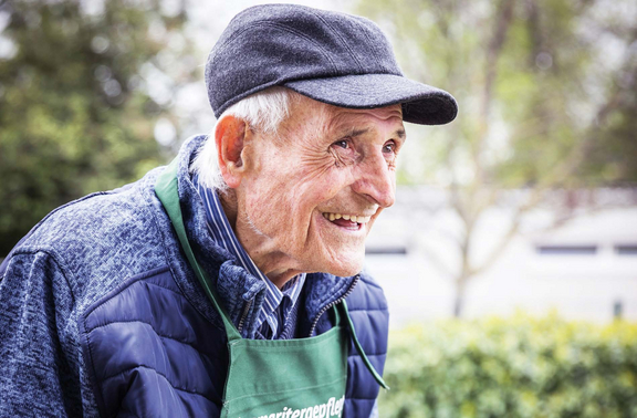 Ein älterer Mann mit einem freundlichen Lächeln trägt eine graue Mütze und eine grüne Schürze. Er steht in einem Garten.