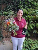 Frau mit Blumenstrauß und Champagnerflasche, lächelt vor grüner Hintergrundpflanzung.
