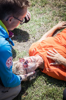 Ein Sanitäter leistet Erste Hilfe einer ältere Frau, die auf dem Boden liegt. Sie trägt ein oranges Oberteil und eine Sonnenbrille. Der Sanitäter beugt sich über sie und scheint ihre Vitalwerte zu überprüfen. Im Hintergrund ist grünes Gras sichtbar.