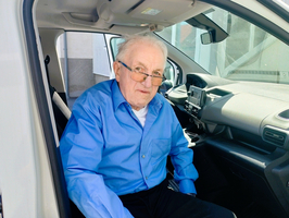 Ein älterer Mann mit Brille sitzt auf dem Fahrersitz eines Fahrzeugs. Er trägt ein hellblaues Hemd und hat graue Haare. Die Inneneinrichtung des Autos ist sichtbar, ebenso ein Teil der Außenumgebung.
