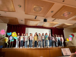 Eine Gruppe von Kindern singt auf einer Bühne vor bunten Papierblumen.