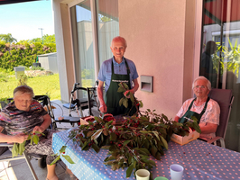 Drei ältere Menschen sitzen an einem Tisch im Freien, umgeben von Pflanzen. Eine Person sortiert Blätter, die anderen lächeln. Sie tragen Schürzen und arbeiten gemeinsam an einem floralen Projekt. Im Hintergrund sieht man einen Garten und Tageslicht, was eine freundliche Atmosphäre schafft.