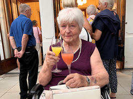 Eine ältere Frau im Rollstuhl hält zwei Cocktails in den Händen.