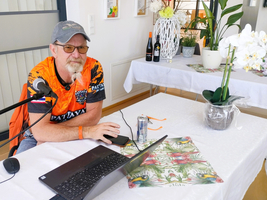 Ein Mann mit Brille und Bart sitzt an einem Tisch mit einem Laptop. Er trägt ein orangefarbenes T-Shirt und hat ein Mikrofon neben sich. Auf dem Tisch stehen eine Getränkedose, ein Notizblock und eine Topfpflanze. Im Hintergrund sind Flaschen und Dekorationen sichtbar.