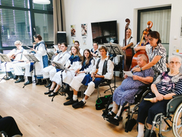 Eine Gruppe von Musikern in traditioneller Kleidung spielt auf einer Bühne. Im Vordergrund sitzen mehrere ältere Menschen in Rollstühlen und schauen aufmerksam zu. Die Atmosphäre ist freundlich und gemeinschaftlich, und die Musiker scheinen Freude an ihrem Auftritt zu haben.