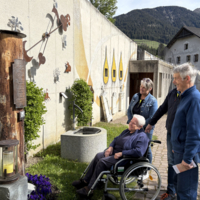 Ältere Frau im Rollstuhl betrachtet mit zwei Begleitpersonen eine Wand mit Kunstwerken und Skulpturen. Die Umgebung ist eine gepflasterte Fläche mit Bäumen im Hintergrund und einem Berg. Die Stimmung ist nachdenklich und freundlich.