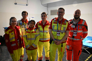 Eine Gruppe von sechs Rettungskräften in leuchtenden gelben und roten Uniformen posiert zusammen in einem Innenraum. Sie wirken freundlich und professionell, bereit für medizinische Einsätze. Im Hintergrund sind medizinische Ausrüstungen sichtbar.