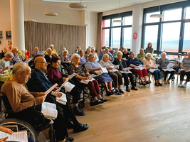 Eine Gruppe älterer Menschen sitzt in einem hellen Raum, der große Fenster hat. Sie halten Liederblätter in den Händen und scheinen an einer gemeinsamen Veranstaltung, wie einem Singen oder einer Aufführung, teilzunehmen.