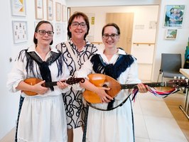 Drei Frauen stehen in einem hellen Raum zusammen. Zwei von ihnen tragen traditionelle Kleidung und halten Musikinstrumente, während die dritte Frau in einem modernen Kleid steht. Alle lächeln in die Kamera. Hintergrund mit gerahmten Dokumenten und Stühlen sichtbar.