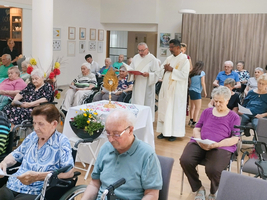 Eine Gruppe älterer Menschen sitzt in einem Raum und hört einem religiösen Akt zu, während zwei Geistliche an einer Tisch mit einer Monstranz stehen. Blumenarrangements sind auf dem Tisch platziert, und einige Anwesende halten Gesangsbücher.