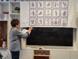 Eine Frau zeigt mit einem Stock auf ein Wandteppich mit symbolischen Bildern.