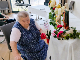 Eine ältere Frau sitzt an einem Tisch mit Blumen und einer Statue.