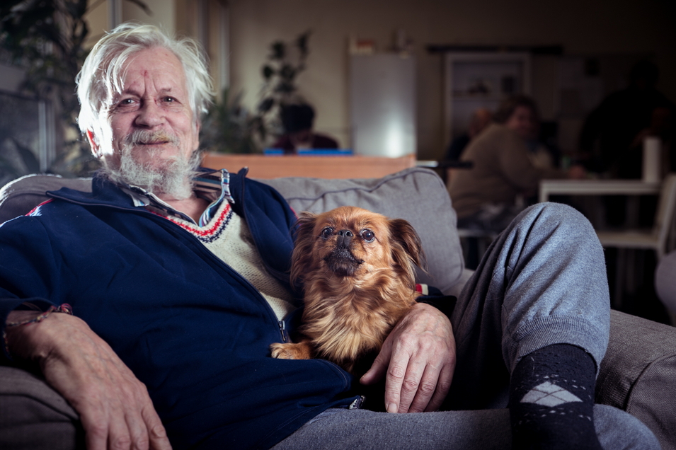 Ein alter Mann sitzt mit seinem Hund auf einem Couchsessel, Obdachlosigkeit, Wohnungslosenhilfe 