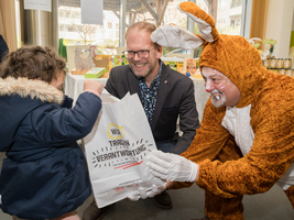 Ein Kind erhält von einem Mann im Hasenkostüm eine Tüte. Der Mann mit Brille lächelt und beobachtet die Übergabe. Im Hintergrund sind Tische mit verschiedenen Produkten und einer festlichen Atmosphäre zu sehen.