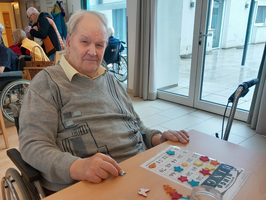 Ein älterer Mann sitzt in einem Rollstuhl an einem Tisch und spielt Bingo. Vor ihm liegt eine Bingo-Karte mit bunten Figuren und Zahlen. Im Hintergrund sind andere Bewohner zu sehen. Der Mann hat graue Haare und trägt einen hellen Pullover.
