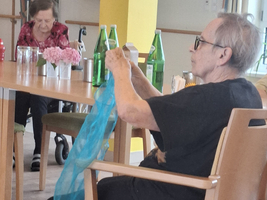 Eine ältere Frau sitzt auf einem Stuhl und hält ein transparentes, blaues Tuch in der Hand. Im Hintergrund sind andere Personen, eine weitere ältere Frau mit einem Rollator und einige Wasserflaschen auf dem Tisch zu sehen.