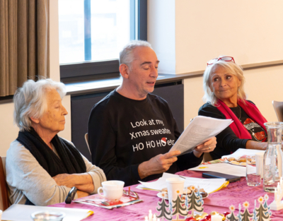 Drei Senioren sitzen an einem schön gedeckten Weihnachtstisch, einer der Personen hält einen Zettel in der Hand und liest etwas vor, Weihnachtsfeiern, Senioren-WGs