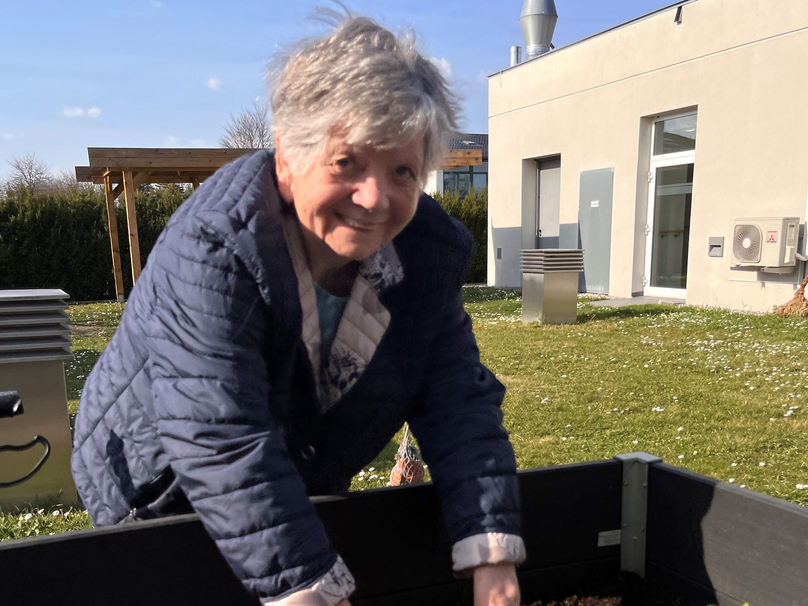 Eine ältere Frau mit grauen Haaren lächelt und arbeitet im Garten. Sie pflanzt Setzlinge in einem Hochbeet mit Erde. Im Hintergrund sieht man ein modernes Gebäude und einen klaren blauen Himmel.