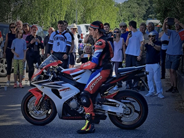 Ein Motorradfahrer in rot-blauer Rennbekleidung sitzt auf einer sportlichen Maschine. Um ihn herum steht eine Gruppe von Zuschauern, die das Geschehen mit Interesse beobachten. Bäume sind im Hintergrund sichtbar, und die Atmosphäre wirkt lebhaft.