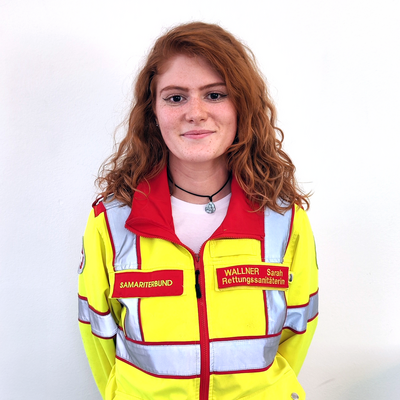 Eine Frau mit roten Haaren trägt eine Rettungsdienstjacke und lächelt.