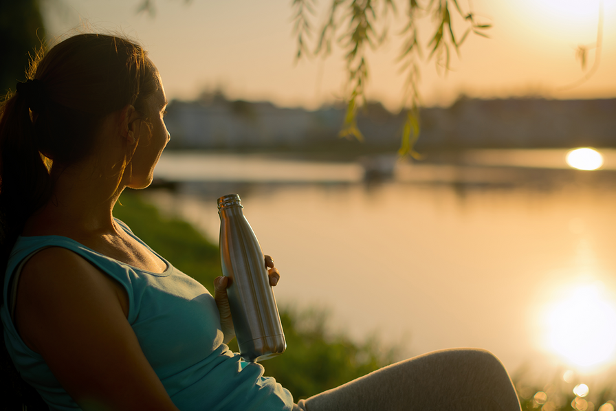 Eine Frau sitzt am Wasser und hält eine Wasserflasche in der Hand.