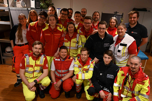 Eine Gruppe von etwa 20 Personen in Rettungsdienst-Uniformen steht in einem Raum. Sie lächeln und posieren zusammen. Im Hintergrund sind Regale und einige technische Ausstattungen zu sehen. Die Mehrheit trägt rot-gelbe Uniformen, während einige in anderen Farben gekleidet sind.