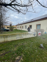 Ein Gebäude mit einem Schild, auf dem „Pflegekompetenzzentrum Strem“ steht. Im Vordergrund steht eine Person, die ein Foto macht. Im Hintergrund sind Fenster und ein leicht bewölkter Himmel zu sehen. Der Bereich ist mit Gras und einigen Blumen gestaltet.