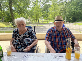 Zwei ältere Menschen sitzen an einem Tisch im Freien, umgeben von Bäumen.