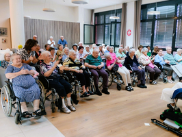 Eine Gruppe älterer Menschen sitzt in einem Raum. Viele nutzen Rollstühle, während sie auf einer Veranstaltung oder Aktivität warten. Einige Personen sind in Gespräche vertieft, während andere entspannt sitzen und lächeln. Der Raum hat große Fenster mit viel natürlichem Licht.